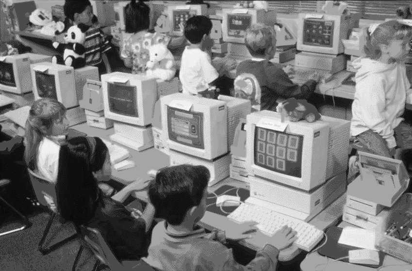 black-and-white photo of kids in a 1990's style computer classroom, one is clearly playing solitaire